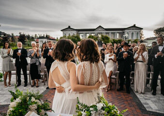 Portrait of two LGBT females lesbians brides posing during wedding ceremony, guests clapping and...