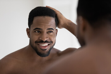 Handsome shirtless muscular build African man standing in bathroom looking in mirror touch his...