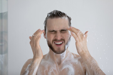Head shot young lathered man takes a shower having eyes tearing up due to sulphate shampoo,...