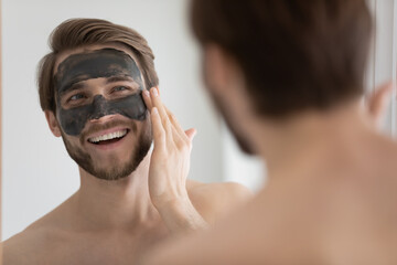 Happy handsome guy looks in mirror apply enriched charcoal facial mask, enjoy face wash procedure,...