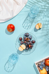 Creative composition made of plate with fruits, glasses, apples on pastel blue sunlit background with white silk cloth and palm tree leaves shadows. Summer and refreshment concept. Harvest theme