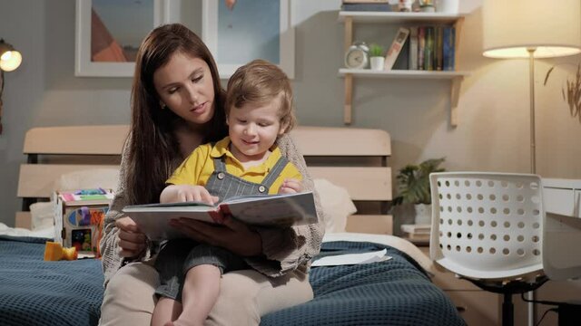 Mom and baby reading. Mom and child 2-3 years old sit in bedroom on bed in evening and read book, woman and boy point at something in book with their finger. Slow motion