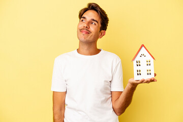 Young mixed race man holding toy house isolated on yellow background dreaming of achieving goals and purposes