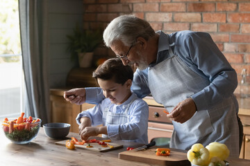 Caring mature grandfather in glasses teaching little grandson to cut vegetables, happy senior grandpa with 8s boy child wearing aprons cooking salad, spending leisure time weekend in kitchen together