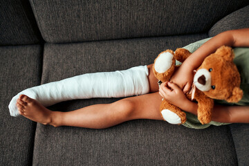 A cute little girl with a broken leg in a cast, sits hugging a teddy bear on the couch at home.