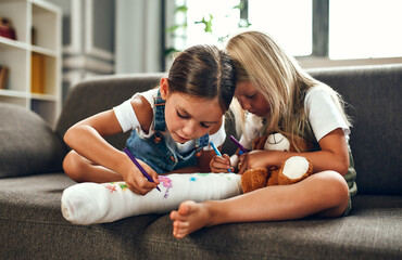 Little girl with a broken leg on the couch. Two sisters draw with felt-tip pens on a plaster bandage. Children have fun and play on the living room couch. - obrazy, fototapety, plakaty