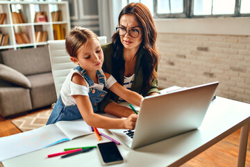 Business woman and mom try to work at the laptop when her little daughter is playing, fooling around and interfering with her. Freelance, work from home.