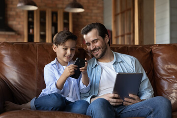 Happy father with little son having fun with electronic devices, relaxing sitting on couch at home, smiling dad holding tablet, 8s boy kid looking at smartphone screen, enjoying weekend together