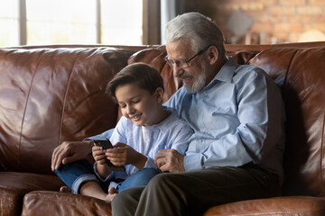 Smiling grandfather with little grandson using phone, sitting on couch, happy grandpa in glasses with 8s boy kid watching funny video or cartoon, playing mobile device game, looking at screen