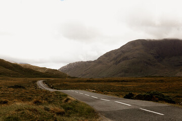 Empty road in Ireland. Autumn in Connemara. Overcast day with clouds, mountains and hills. Brown dried grass in the background.	