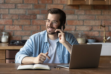 Fototapeta na wymiar Smiling man in glasses talking on smartphone with friend or colleague, sitting at wooden table with laptop at home, friendly businessman freelancer advisor chatting, consulting client by phone call