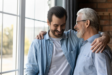 Fototapeta na wymiar Close up happy mature father and grownup son hugging cuddling enjoying tender moment, standing at home, smiling young man with senior dad in glasses having fun, two generations good relationship