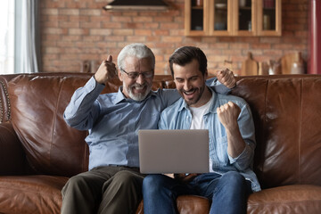 Overjoyed young man and mature father celebrating success, using laptop, looking at device screen,...