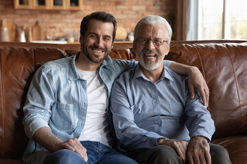 Family portrait smiling mature father and grownup son hugging, sitting on couch at home, happy...