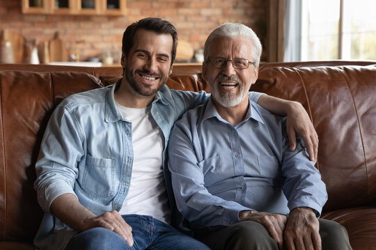 Family portrait smiling mature father and grownup son hugging, sitting on couch at home, happy young man with senior dad looking at camera, posing for picture, enjoying weekend together