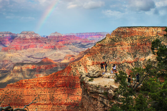 Tourists On Outcropping Of Grand Canyon With Phones On Selfie Sticks - South Rim Arizona With Geological Layers Stretching To Horizon Behind Them And Rainbow In Sky