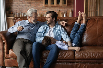Happy three generations of men relaxing on couch together, sitting enjoying lazy leisure time at...