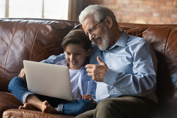 Happy senior grandfather with little grandson using laptop together, sitting on couch at home, smiling mature grandpa and 8s boy kid making video call, chatting online, looking at computer screen