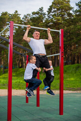 Fototapeta premium Caucasian man and two boys doing exercises outdoors. The father pulls himself up on the horizontal bar with his sons on the playground.