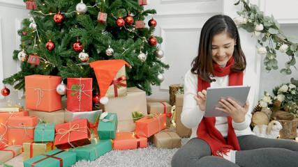 Asian woman using smart tablet sit at christmas tree xmas present gift box in cozy living room....