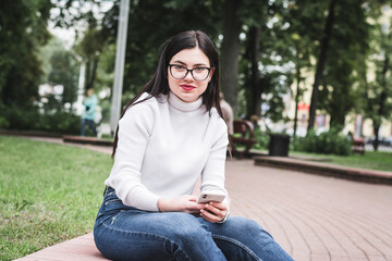 Beautiful stylish girl in jeans and a white sweater outdoors in the park with a phone