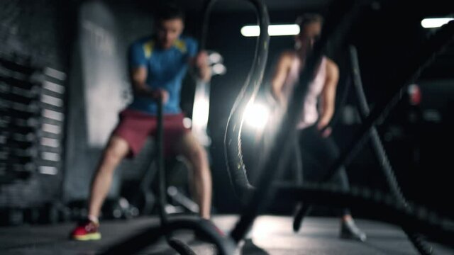 A fitness athletes are training using battle ropes in the dark gym. Friends are doing exercises bodybuilding indoors. Intense sport workout. Concept of lifestyle fitness health and sports.