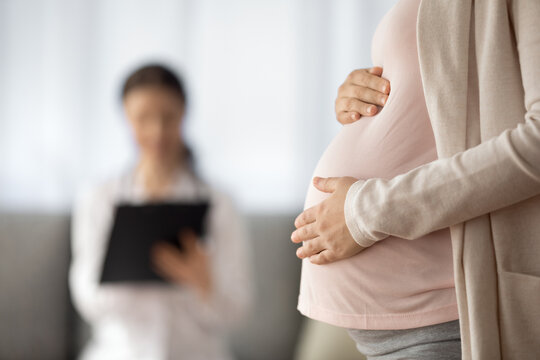 Preparing For Baby Birth. Close Up Of Woman Expecting Baby Having Appointment With Doctor At Antenatal Clinic Prenatal Healthcare Center. Focus On Young Female Holding Hands On Baby Bump In Doc Office