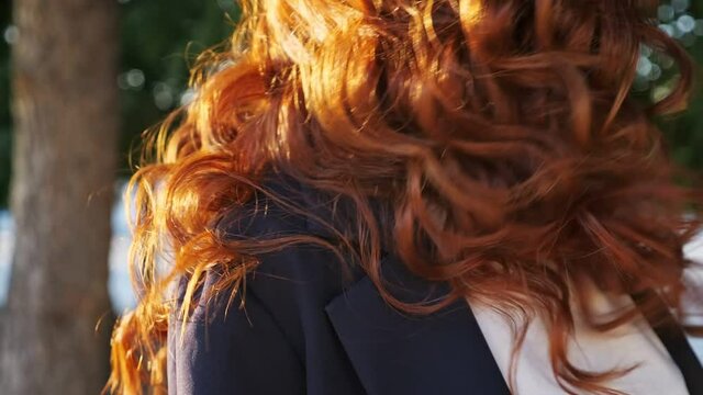 Young woman in jacket shakes long loose curly copper hair lit by sunset light standing on city street with green trees close view slow motion