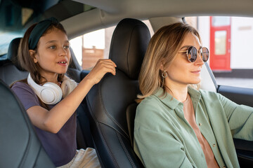 Cute little girl with headphones holding by black leather seat of her mom driving electric car
