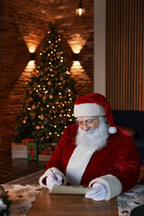 Content senior Santa Claus in red costume sitting at table in living room with Christmas tree and signing letter