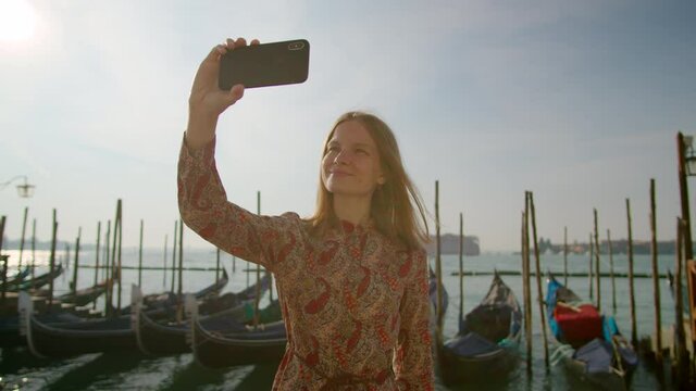 Beautiful Woman Takes Selfie Photos by Phone with Gondolas in Venice Grand Canal, Italy, Europe. Young Lady Tourist Travels alone at Pandemic. Blogging and Landmark Sightseeing Concept. 4K orbit shot