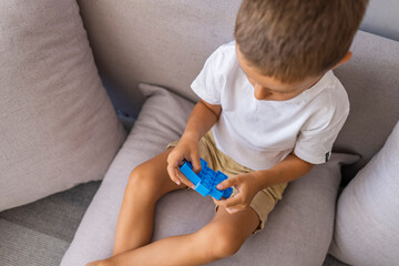 A small child is sitting on the sofa and holding a pop it toy. Portrait of concentrated cute little boy playing popit toy. Antistress toy for children. Leisure for kids. 