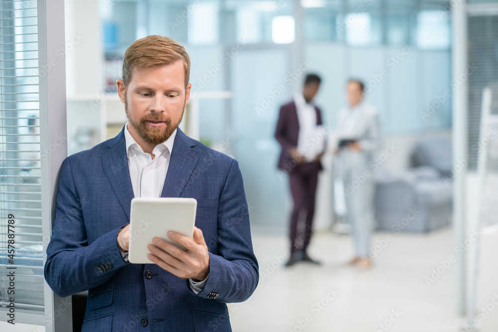 Poster serious business leader scrolling through online data in tablet against his colleagues in office