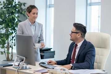 Busy mature employer looking at his secretary in formalwear carrying him cup of tea