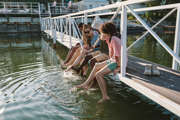 Cheerful young intercultural friends splashing water by legs while sitting in row along wooden bridge