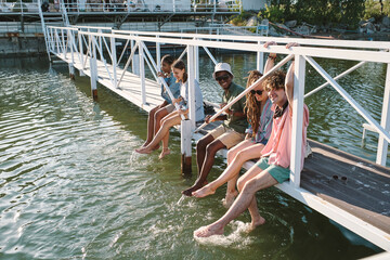 Group of young carefree friends relaxing on bridge and splashing water on hot summer day