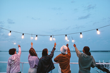Back view of young ecstatic friends with bengal lights enjoying summer evening by lake