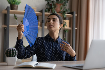 Overheated tired Indian businesswoman waving paper fan at home office workplace, suffering from...