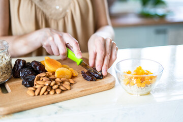 female hands cut dried fruits and nuts with a green knife: dried apricots, almonds and dates on a wooden cutting board into a glass bowl