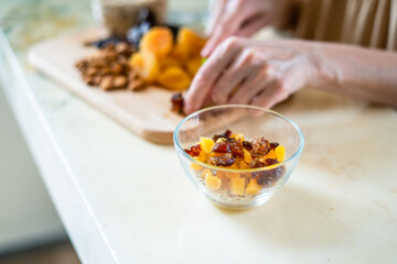 female hands preparing a healthy breakfast, cut dried fruits and nuts with a green knife: dried apricots, almonds and dates on a wooden cutting board into a glass bowl