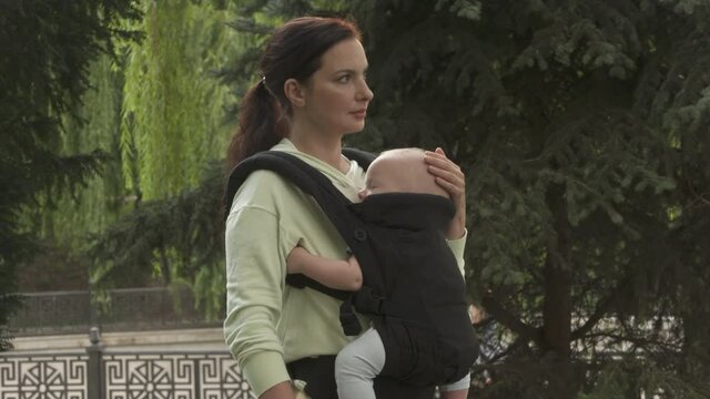 Mother with baby boy in infant kid carrier sling on a walk outdoors in city park. High quality 4k footage
