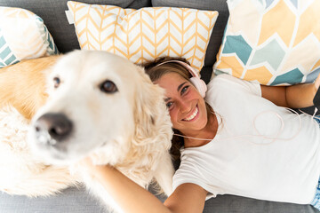 Selective focus of caucasian blonde woman playing with dog in the couch at home. Top view of caucasian woman listening to music with headphones playing with dog. People and dogs lifestyles.