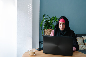 Young muslim woman wearing a hijab using technology indoors. Horizontal panoramic view of arabic woman working with laptop at home. Technology and muslim women lifestyle.