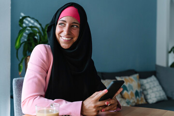 Young muslim woman wearing a hijab using technology indoors. Horizontal side view of arabic woman working with smartphone at home. Technology and muslim women lifestyle.