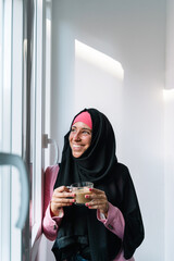 Young muslim woman wearing a hijab drinking coffee indoors. Vertical side view of arabic woman relaxing next to a window at home. Muslim women lifestyle indoors.