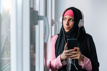 Young muslim woman wearing a hijab using technology indoors. Horizontal side view of arabic woman listening to music with smartphone next to a window at home. Technology and muslim women lifestyle.