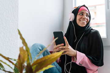 Cheerful muslim woman wearing a hijab using smartphone indoors. Horizontal side view of arabic woman listening to music with headphones next to a window at home. Technology and muslim women lifestyle