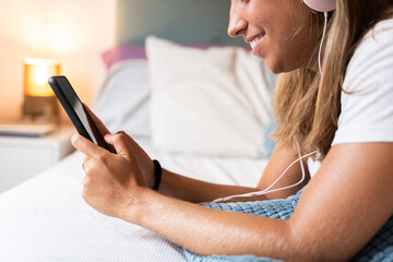 Obraz na płótnie Canvas Unrecognizable woman using smartphone indoors. Horizontal side view of caucasian woman listening to music with headphones lying in bed at home. Technology and women lifestyle indoors