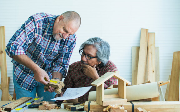 Two senior mixed race diverse happy retired old male woodworkers or carpenters wearing check shirts, creating DIY wooden car toy together for decoration and furniture. Hobby, Retirement Concept.