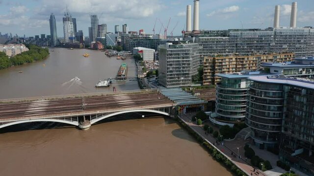 Forwards fly down to multitrack railway bridge. Modern apartment buildings on river bank. Tall modern skyscrapers in background. London, UK